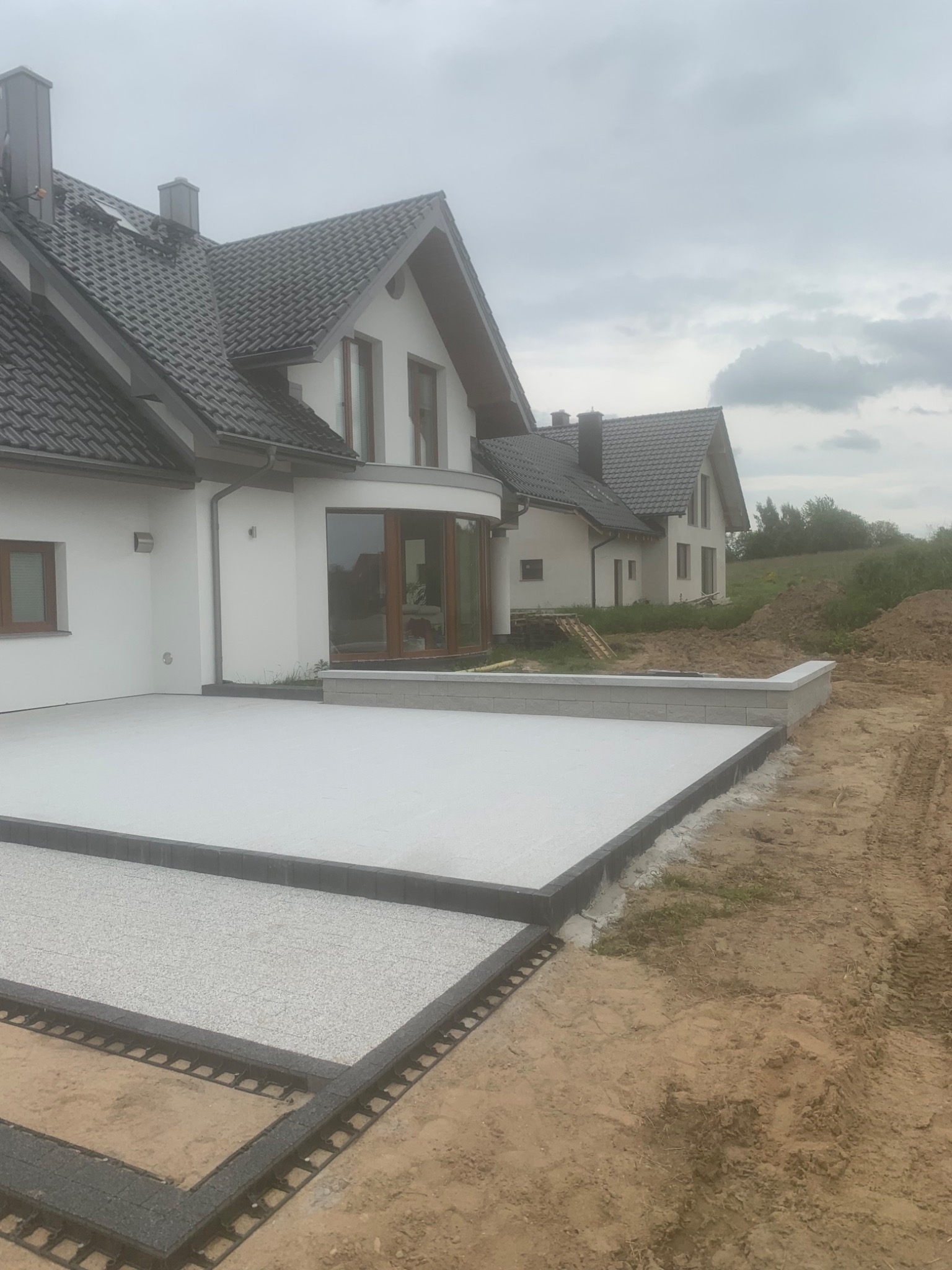 Nowoczesny taras z jasnoszarej nawierzchni żywicznej obramowany ciemnoszarą kostką, widok na dom jednorodzinny z grafitową dachówką i przygotowany teren pod ogród.