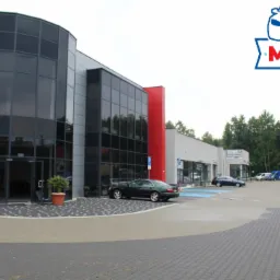 Nowoczesny budynek warsztatu samochodowego z przeszkloną fasadą, zaparkowanymi samochodami i logo firmy w prawym górnym rogu.