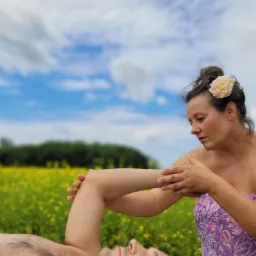 Kobieta z różą we włosach wykonuje masaż ramienia mężczyzny leżącego na tle łąki i błękitnego nieba z chmurami.