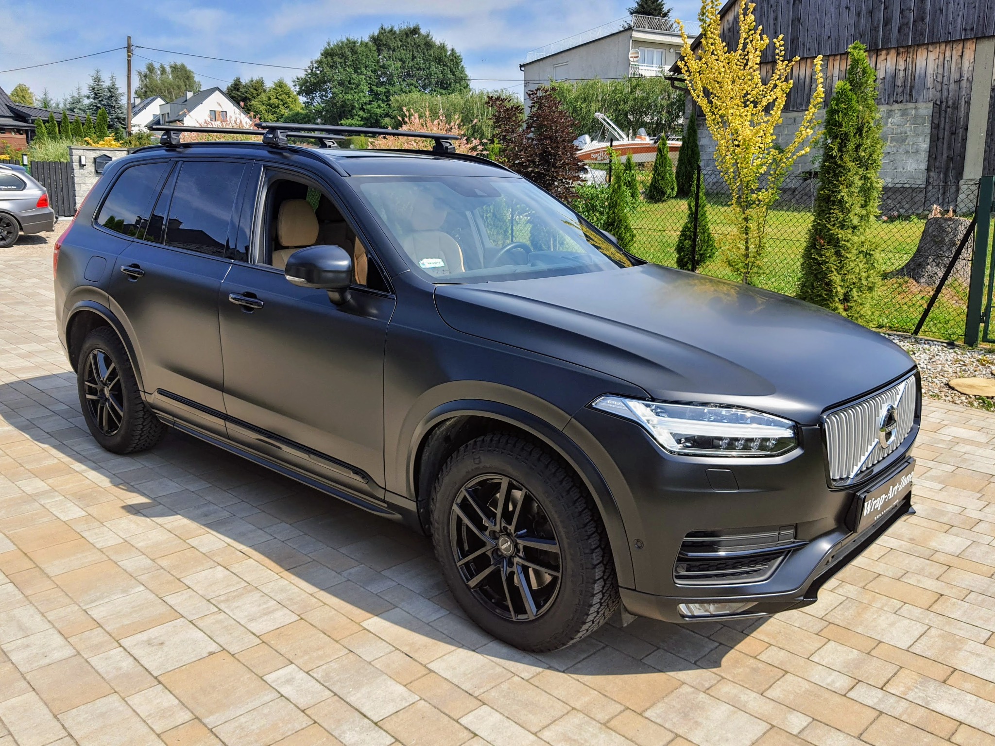 Czarny matowy SUV Volvo XC90 z relingami dachowymi na kostce brukowej, widoczny z boku, z logo firmy Wrap-Art-Zone na przednim zderzaku.