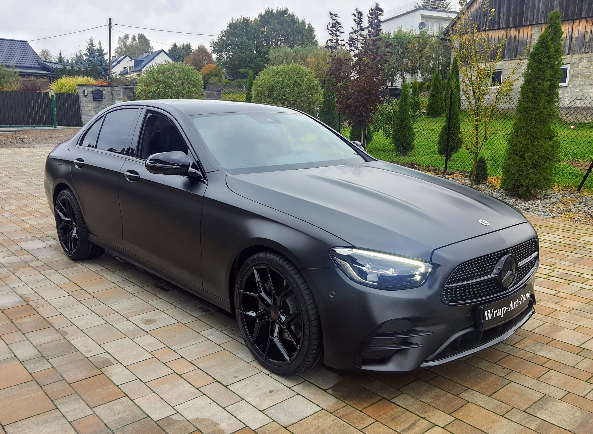 Matowy, czarny Mercedes-Benz E-Klasa z czarnymi felgami, zaparkowany na kostce brukowej przed domem.