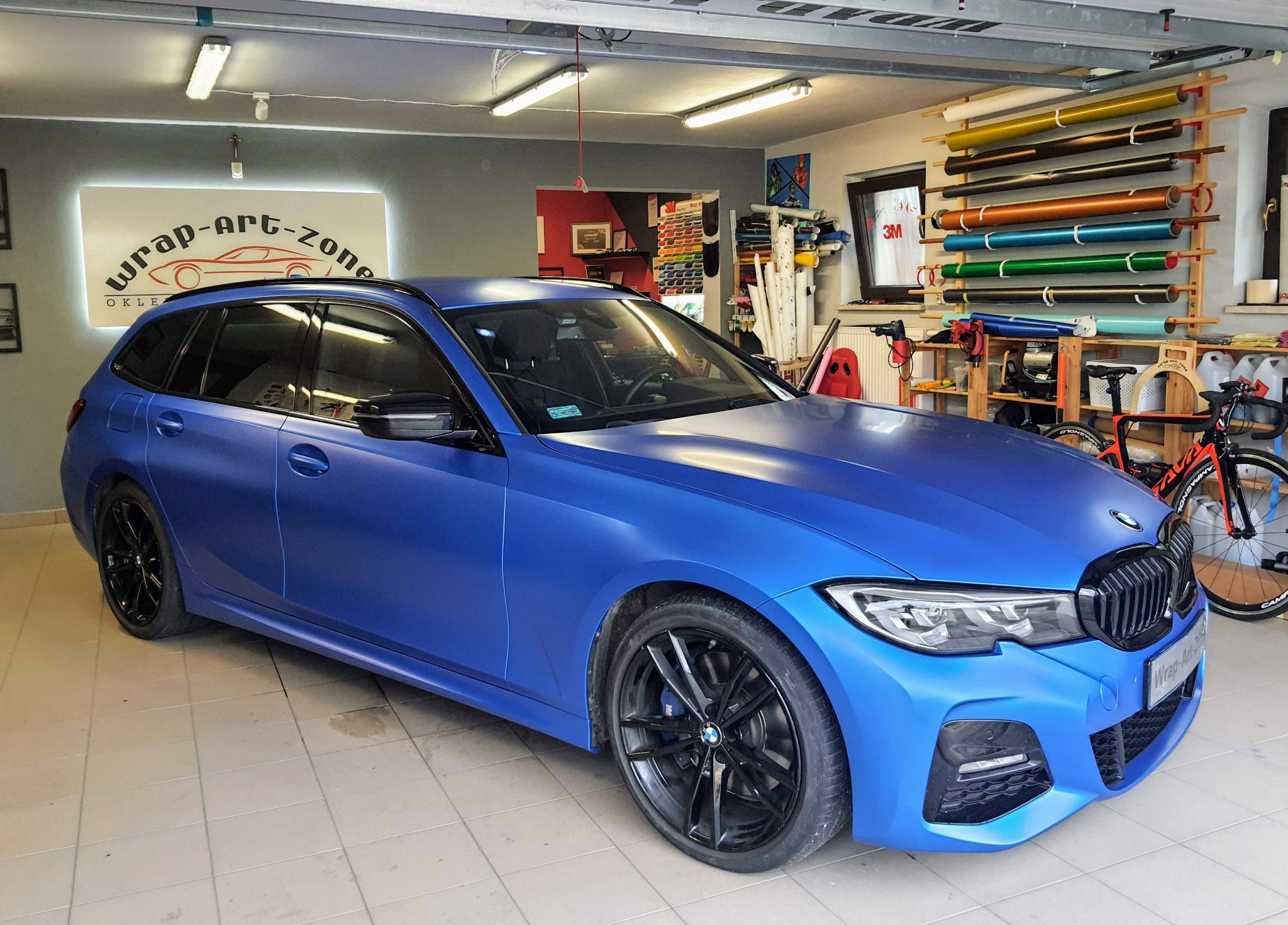Niebieskie BMW kombi z matową folią w warsztacie, widoczne rolki folii na ścianie i logo firmy Wrap-Art-Zone.