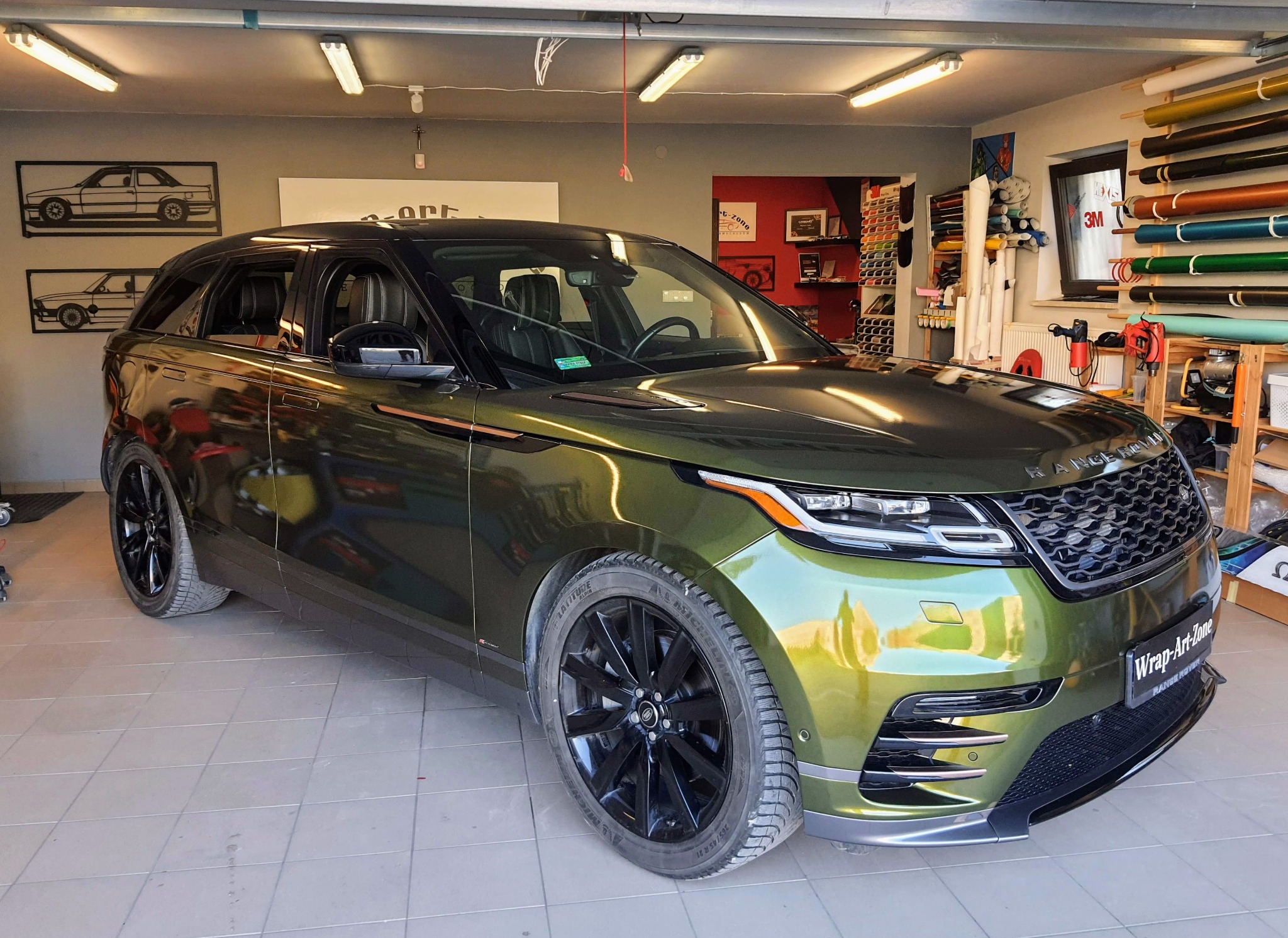 Range Rover Velar w zielonej folii z połyskiem, zaparkowany w garażu z widocznymi rolkami folii i narzędziami, z logo 'Wrap-Art-Zone' na przednim zderzaku.