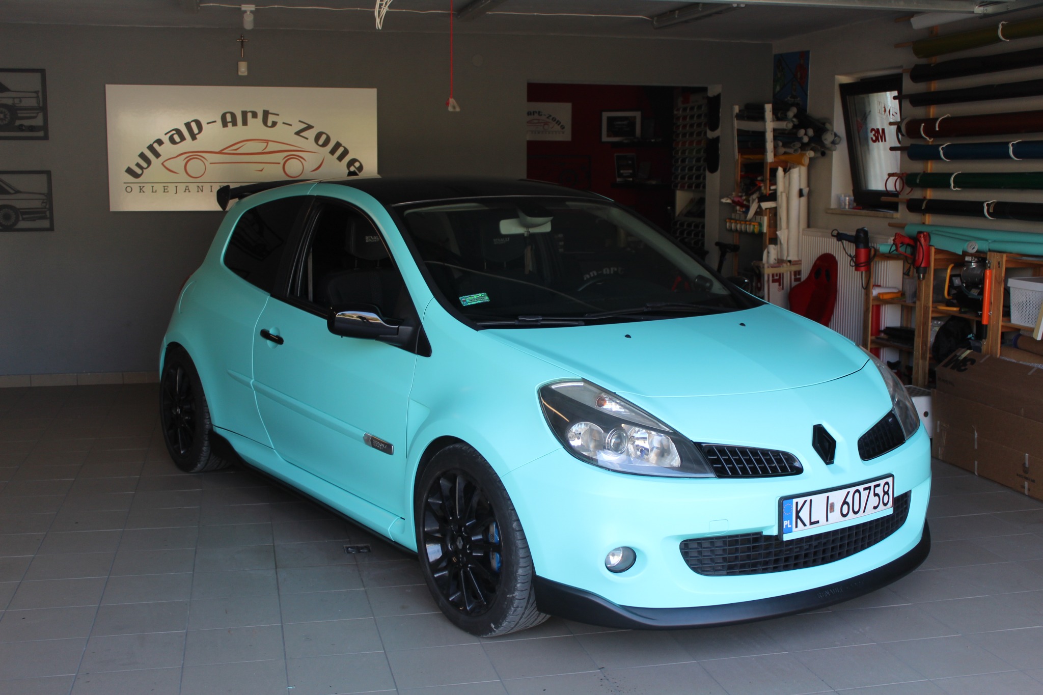 Renault Clio w kolorze miętowym z czarnym dachem i felgami, zaparkowany w garażu z widocznym logo firmy oklejającej pojazdy 'wrap-art-zone' w tle.