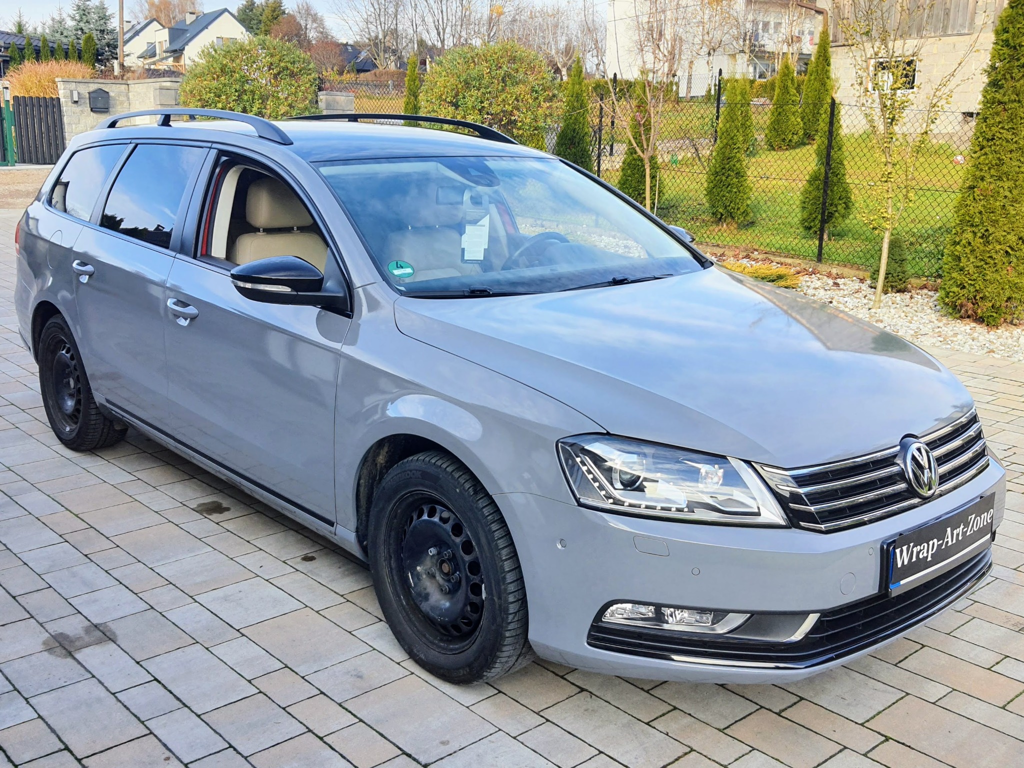 Szary samochód kombi z czarnymi felgami stalowymi i logo firmy Wrap-Art-Zone na przednim zderzaku, zaparkowany na kostce brukowej przed domem.