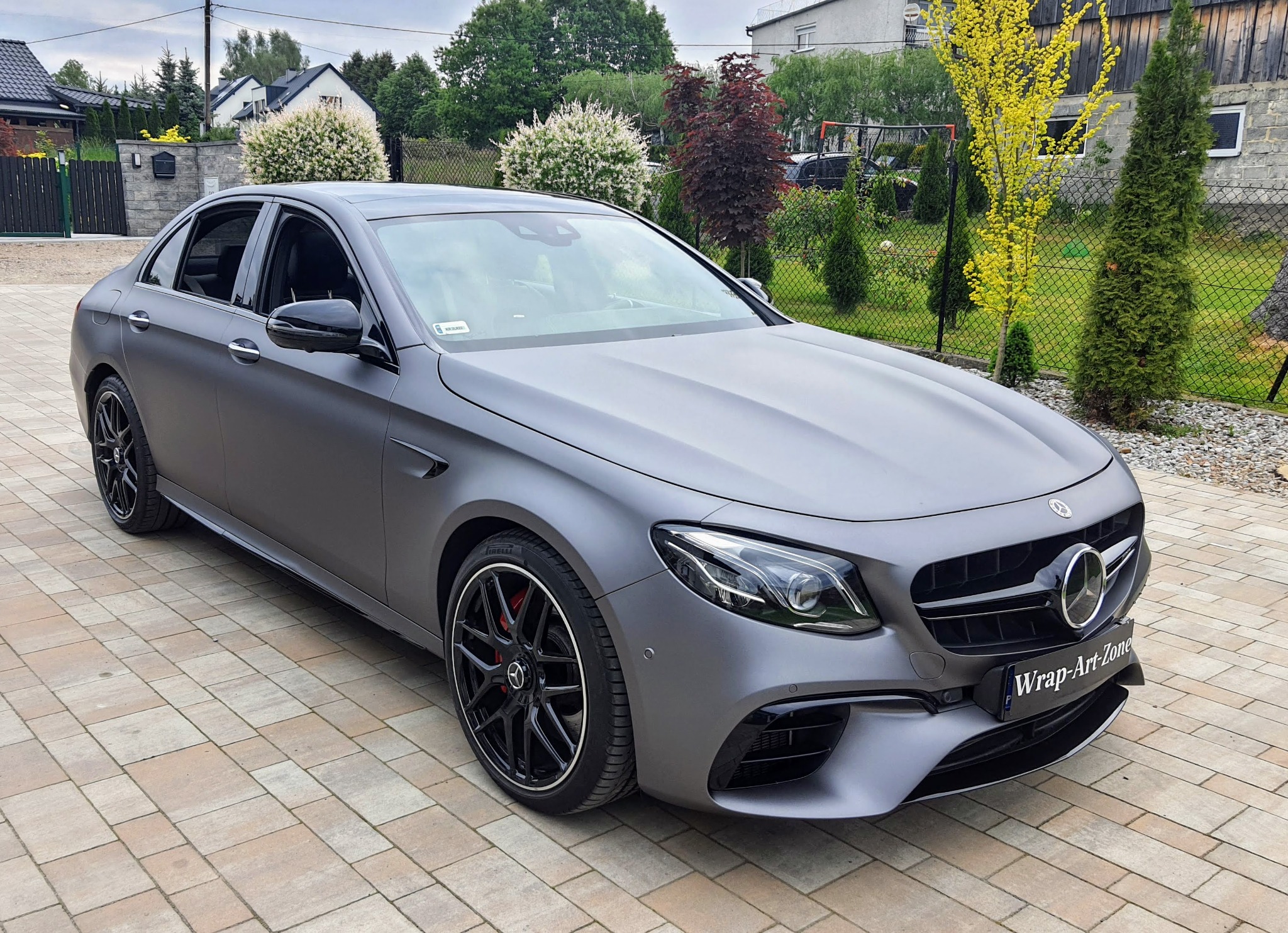 Matowy, szary Mercedes-Benz E63 AMG z czarnymi felgami na kostce brukowej przed domem z zadbanym ogrodem. Na przednim zderzaku tabliczka 'Wrap-Art-Zone'.