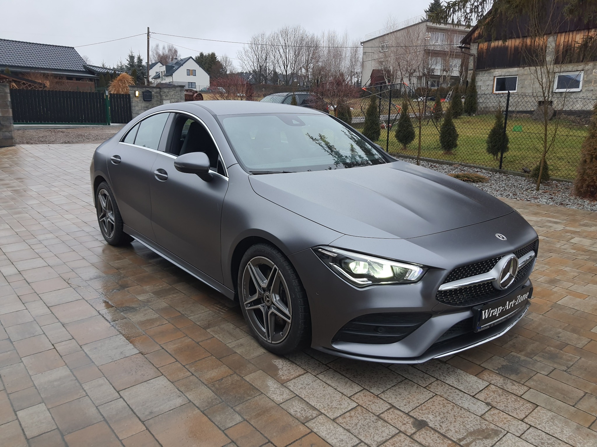 Matowy, szary Mercedes-Benz CLA po zmianie koloru folią, zaparkowany na brukowanym podjeździe przed domem.