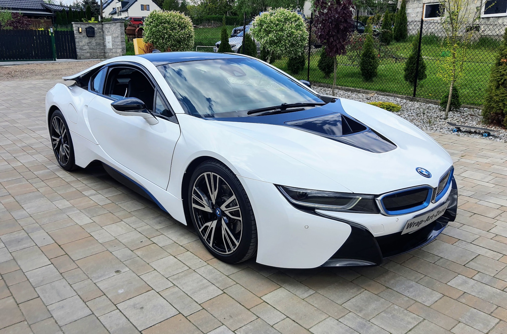 Białe BMW i8 z czarnym dachem zaparkowane na kostce brukowej, widok z boku, na przednim zderzaku naklejka z nazwą firmy.