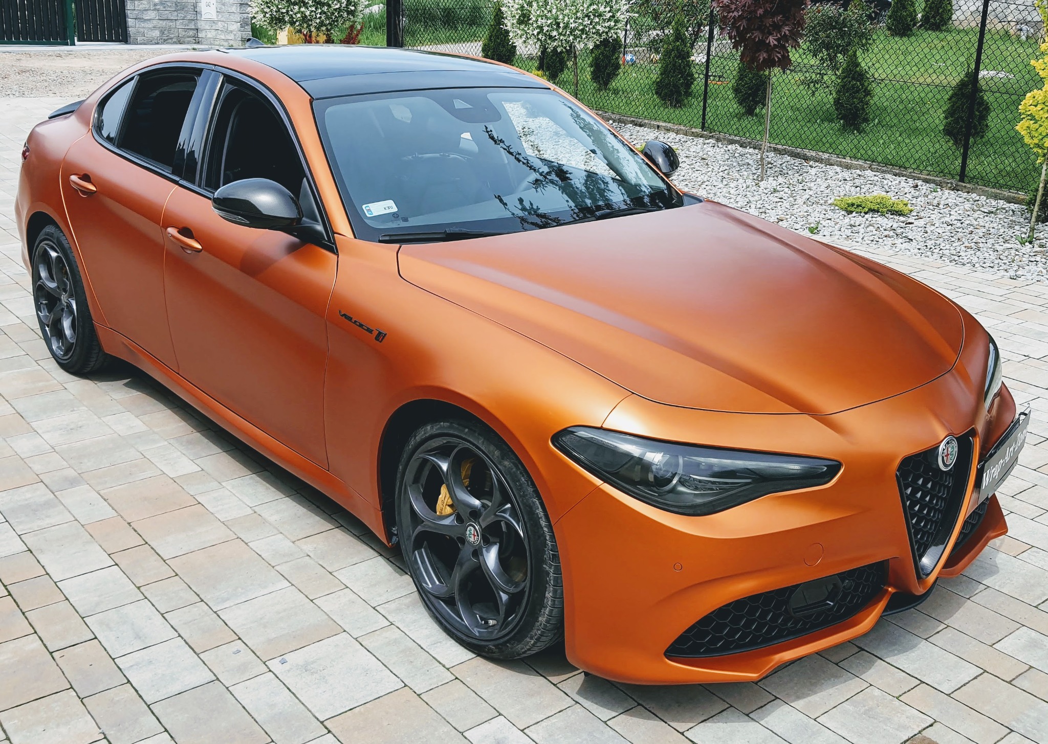 Alfa Romeo Giulia Veloce w matowym, pomarańczowym oplocie karoserii z czarnym dachem, zaparkowana na kostce brukowej.