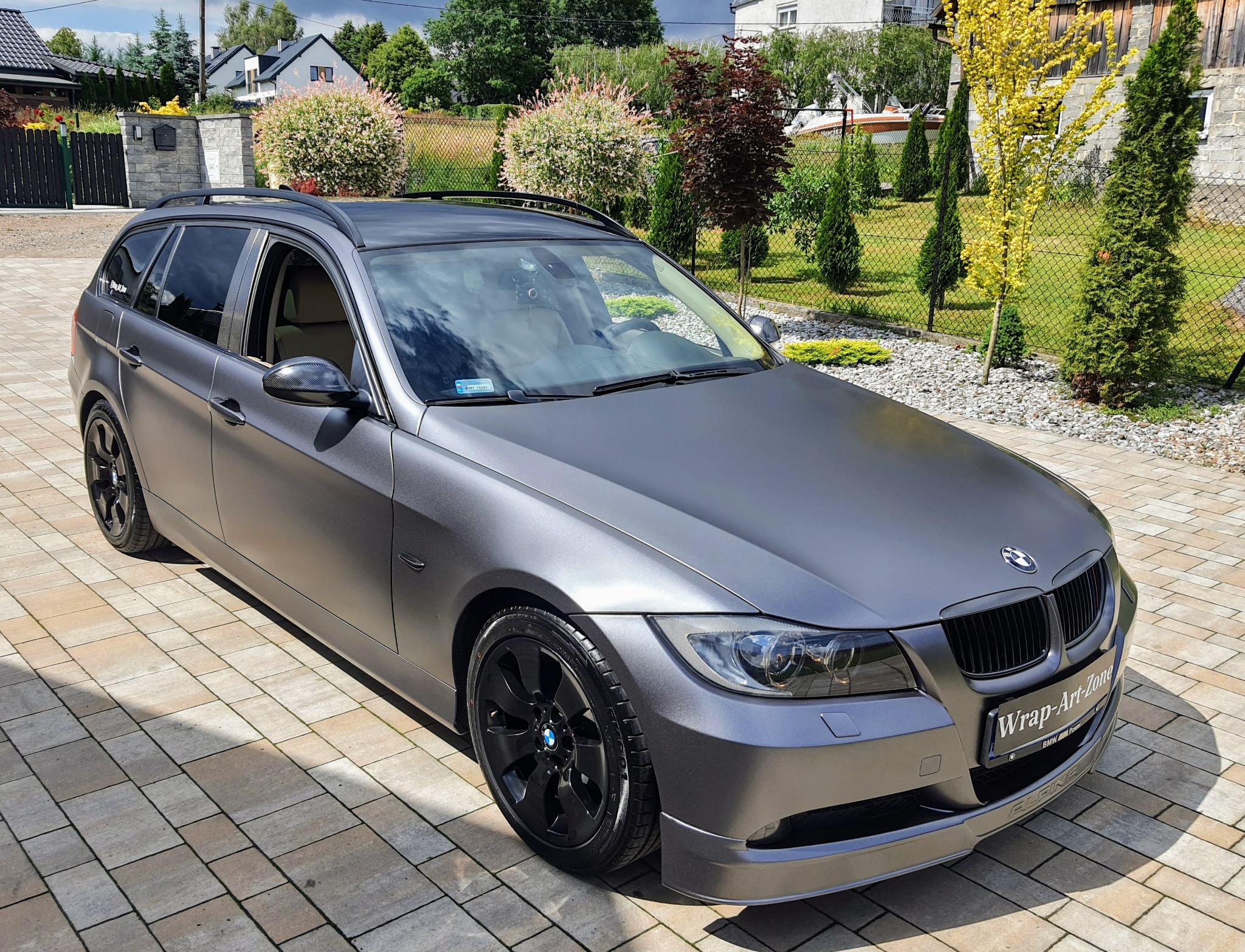 Grafitowe BMW serii 3 kombi po tuningu, z matową folią i czarnymi felgami, zaparkowane na kostce brukowej przed zadbanym ogrodem.