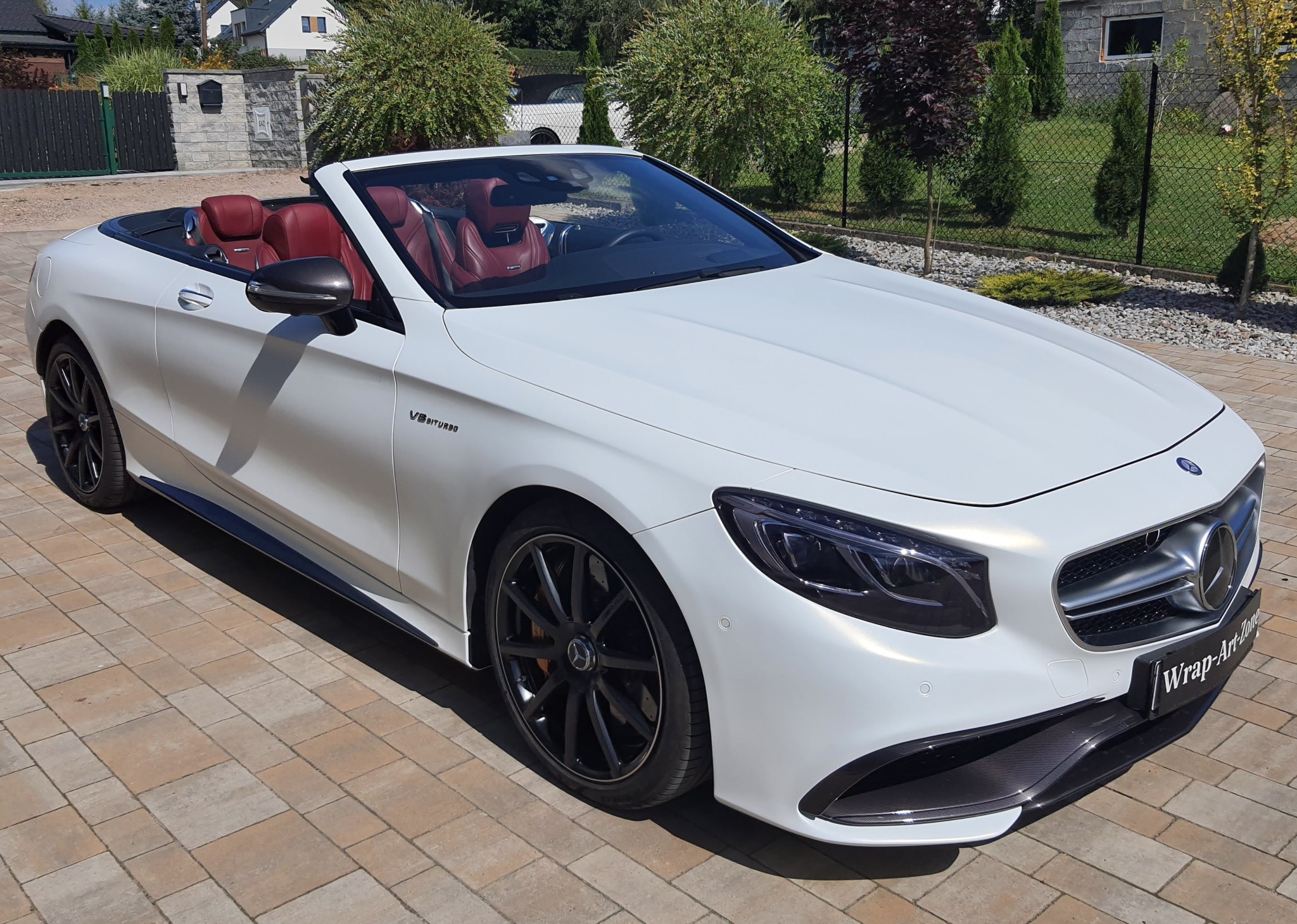 Biały kabriolet Mercedes-Benz S63 AMG z czerwonymi skórzanymi siedzeniami i karbonowymi elementami, zaparkowany na kostce brukowej z widocznym logo Wrap-Art-Zone.