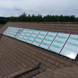 Panele słoneczne marki Vaillant z folią ochronną, zamontowane na brązowym, spadzistym dachu z dachówki ceramicznej, widok z góry na tle zieleni i pochmurnego nieba.