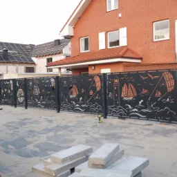 Czarna, metalowa brama wjazdowa z wycinanym wzorem motywów morskich: żaglówki, latarnie, delfiny, kompas i fale, zainstalowana przed domem z czerwonej cegły i budynkiem w trakcie budowy.