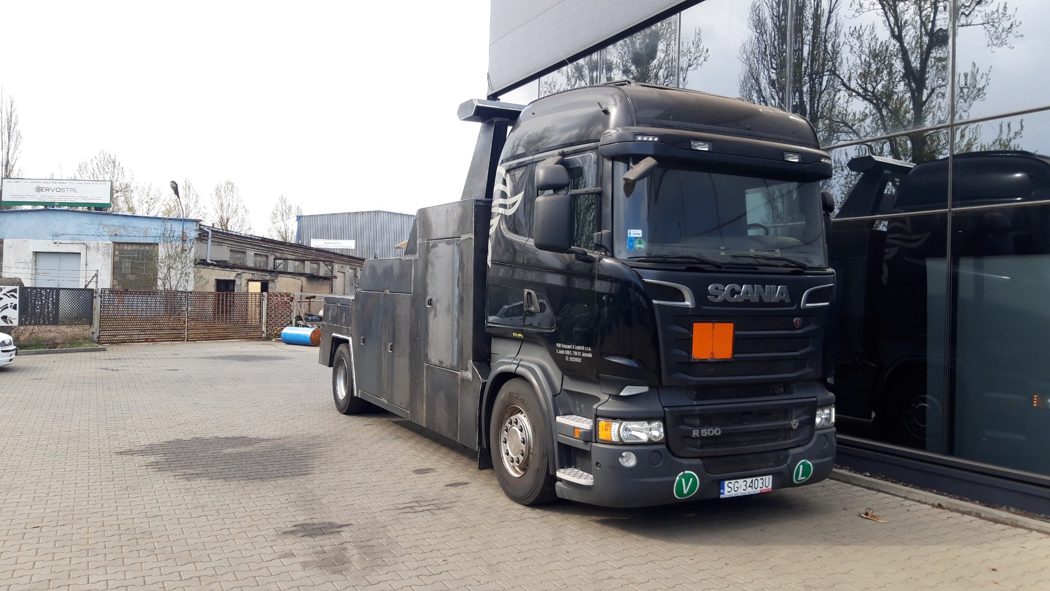 Czarna laweta Scania R500 zaparkowana na brukowanym placu przed budynkiem z szklaną fasadą, z widocznym znakiem firmy SERVOSTAL w tle.