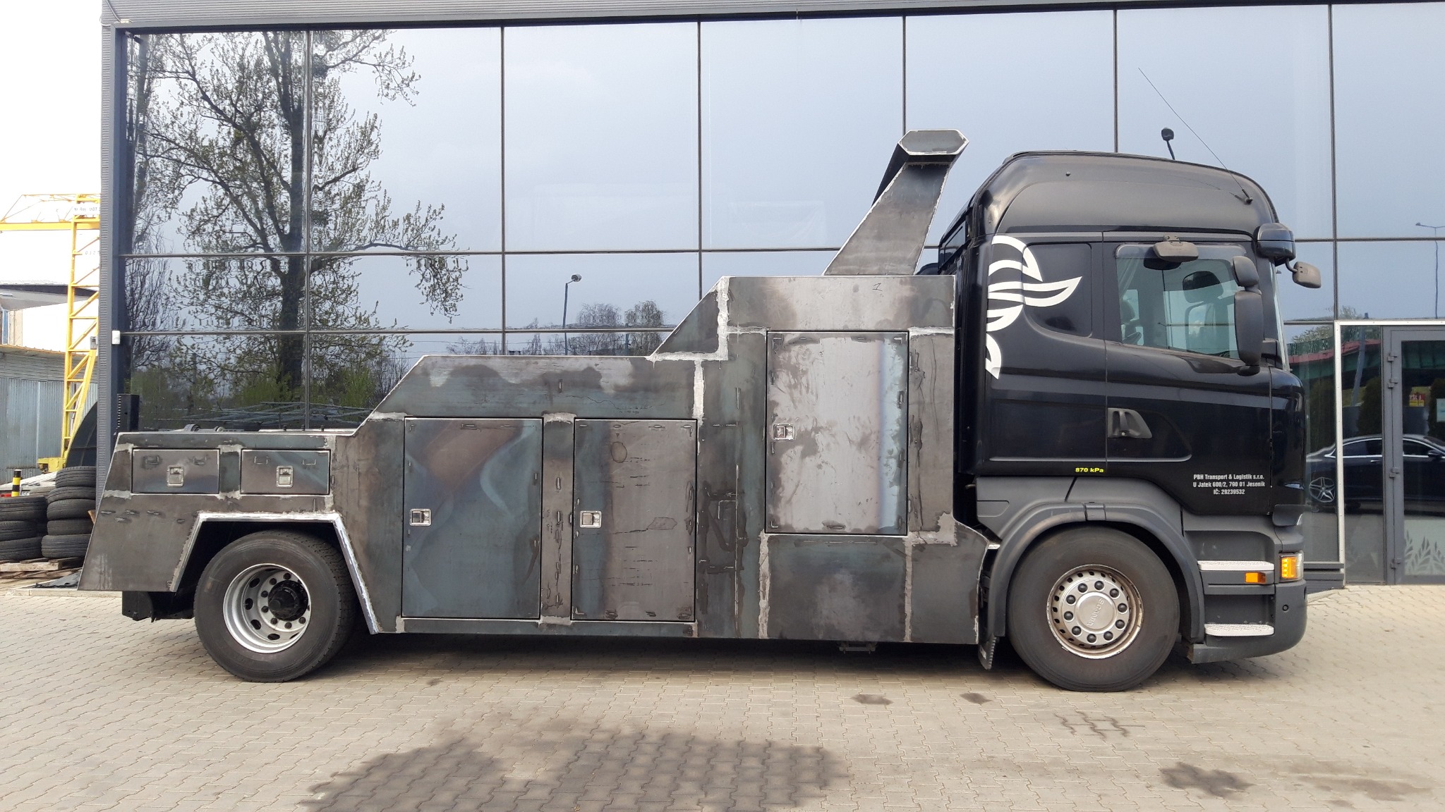 Czarny samochód ciężarowy Scania z niestandardową, surową konstrukcją z metalowych płyt, przypominającą zabudowę specjalną, zaparkowany na kostce brukowej przed szklanym budynkiem. Widoczne spawane...