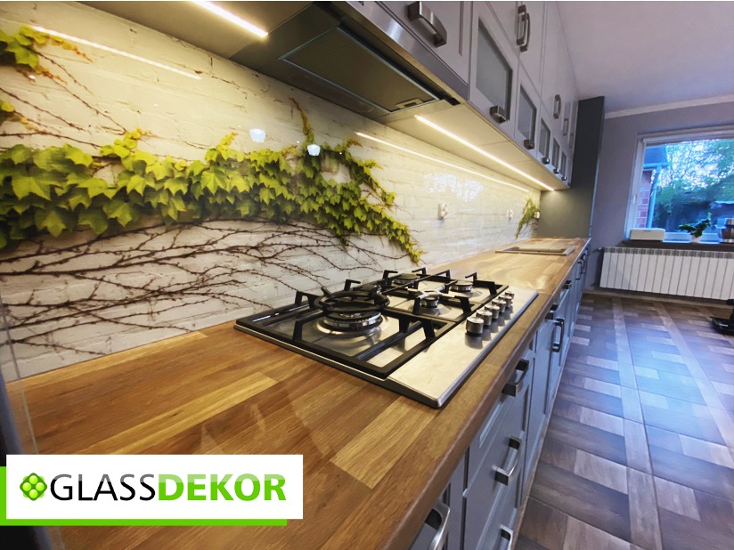 Szklany panel kuchenny z motywem pnącej się roślinności, podświetlony listwą LED, zamontowany nad drewnianym blatem i płytą gazową. Widoczne szare szafki z metalowymi uchwytami oraz logo firmy...