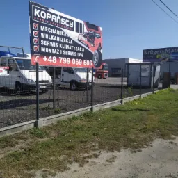 Billboard reklamowy serwisu samochodowego Kopańscy z ofertą usług mechaniki pojazdowej, wulkanizacji, serwisu klimatyzacji oraz montażu i serwisu LPG, na tle zaparkowanych samochodów dostawczych...
