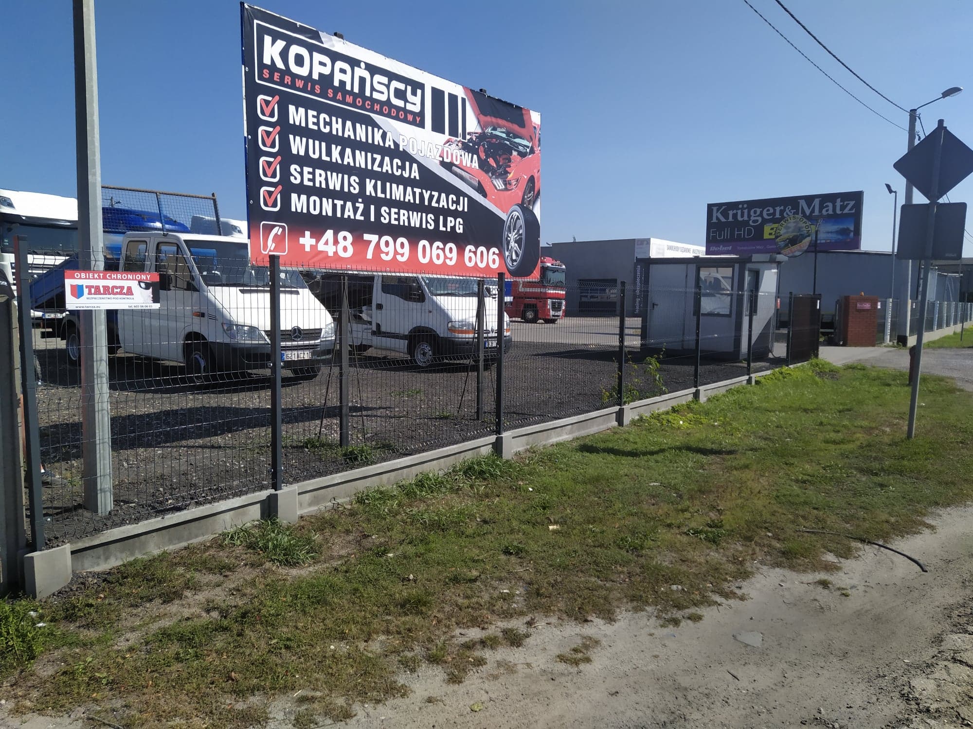 Billboard reklamowy serwisu samochodowego Kopańscy z ofertą usług mechaniki pojazdowej, wulkanizacji, serwisu klimatyzacji oraz montażu i serwisu LPG, na tle zaparkowanych samochodów dostawczych...