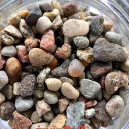 Różnokolorowe, drobne kamienie w szklanym pojemniku, widoczne z góry, ukazujące zróżnicowaną teksturę i odcienie szarości, brązu i czerwieni.