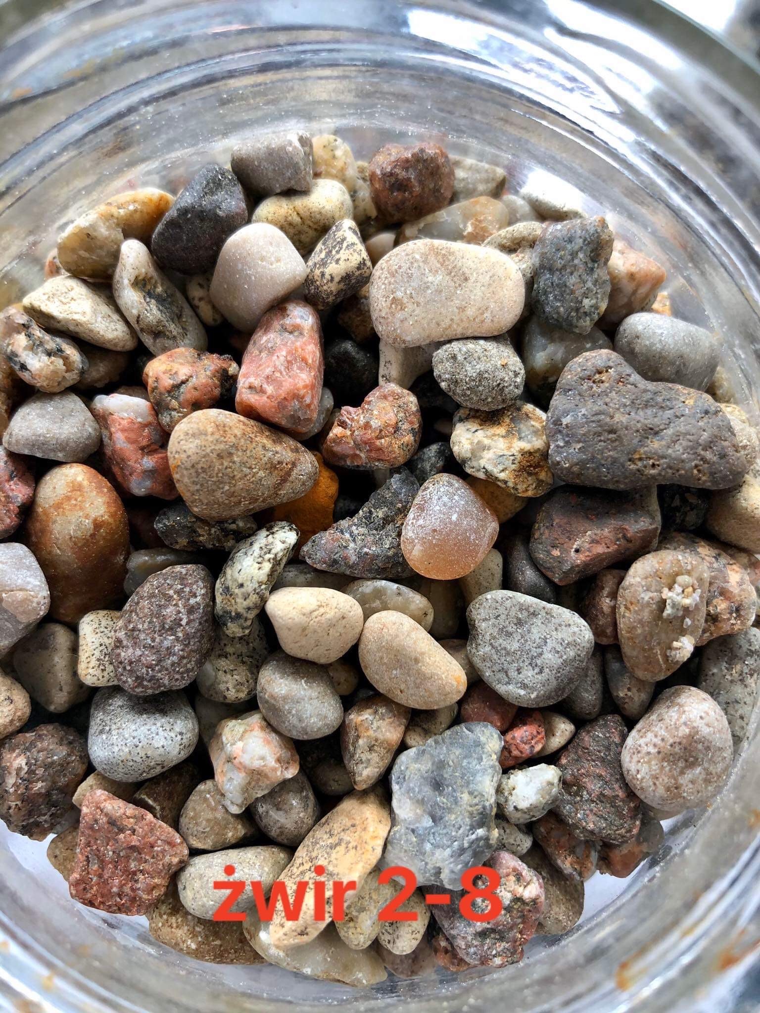 Różnokolorowe, drobne kamienie w szklanym pojemniku, widoczne z góry, ukazujące zróżnicowaną teksturę i odcienie szarości, brązu i czerwieni.