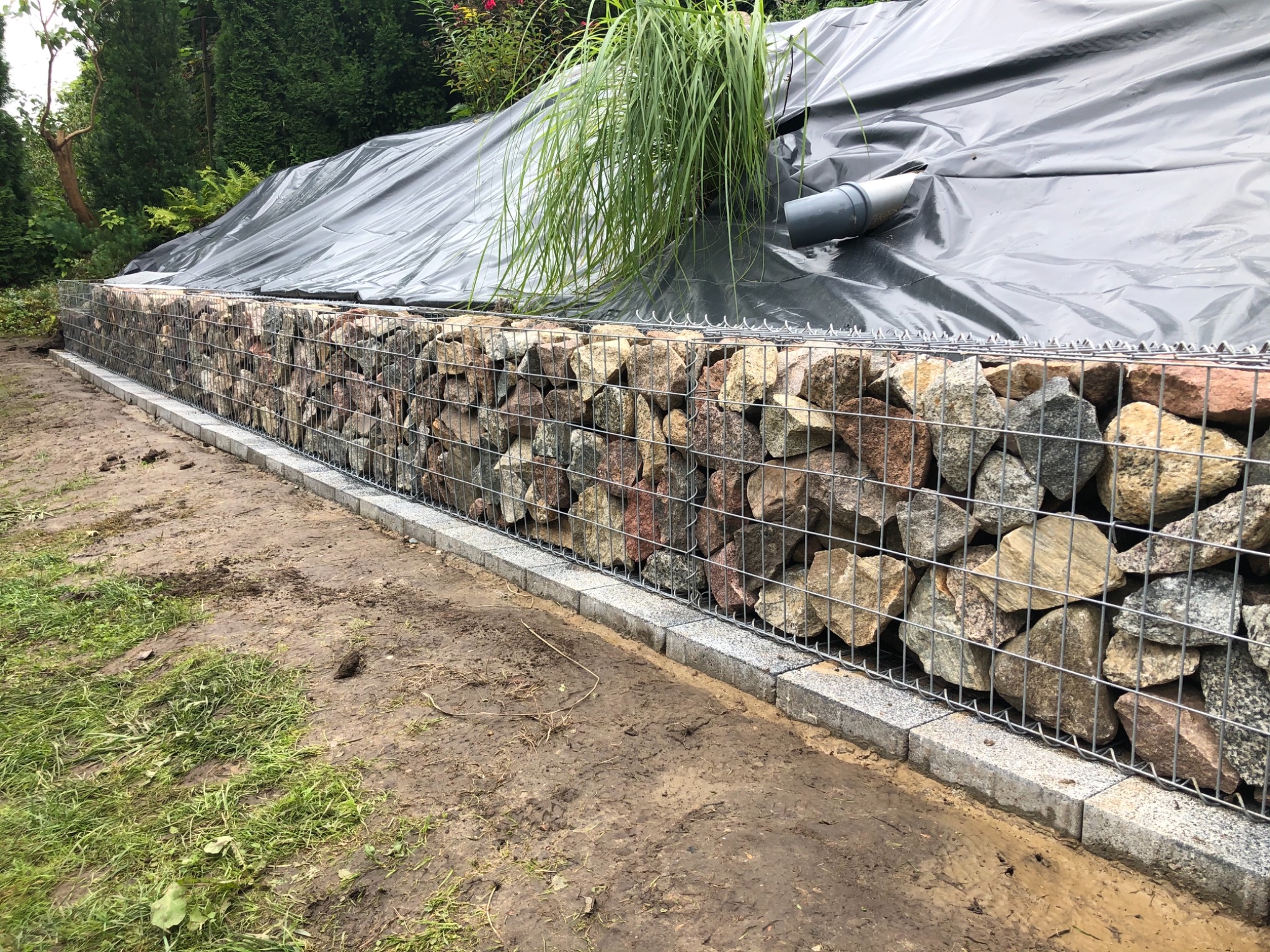 Gabion z kamieniami umocniony obrzeżem z kostki granitowej, zabezpieczony folią, z widoczną rurą drenażową i roślinnością w tle.