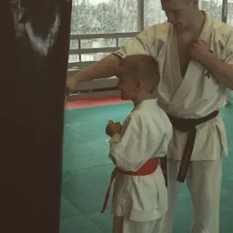 Instruktor w białym kimonie z czarnym pasem uczy chłopca w białym kimonie z czerwonym pasem uderzać w worek treningowy na tle zielonej maty i okna.