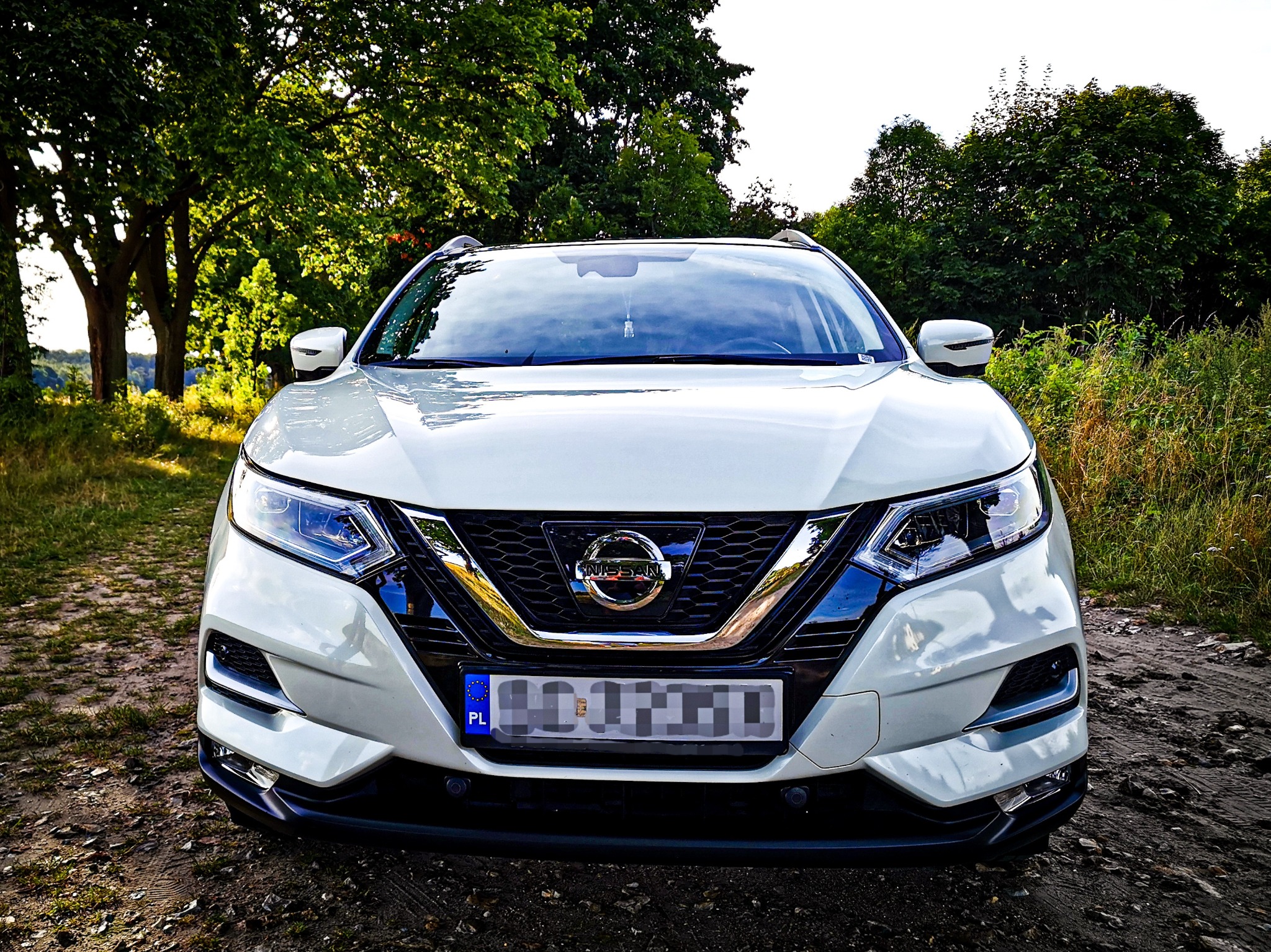 Biały samochód marki Nissan, widok z przodu, zaparkowany na polnej drodze w otoczeniu zieleni.