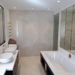 Nowoczesna łazienka z dwoma umywalkami nablatowymi, lustrem w czarnej ramie, marmurowymi płytkami ściennymi i wanną z obudową w kolorze czerni i bieli, oraz złotą armaturą.