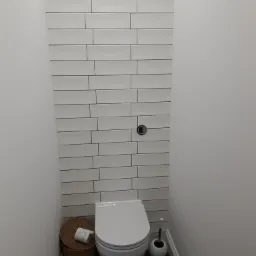 Wąska toaleta z białymi, poziomymi płytkami na ścianie, wiszącą miską WC, koszem wiklinowym na papier toaletowy i wzorzystą podłogą w odcieniach szarości.