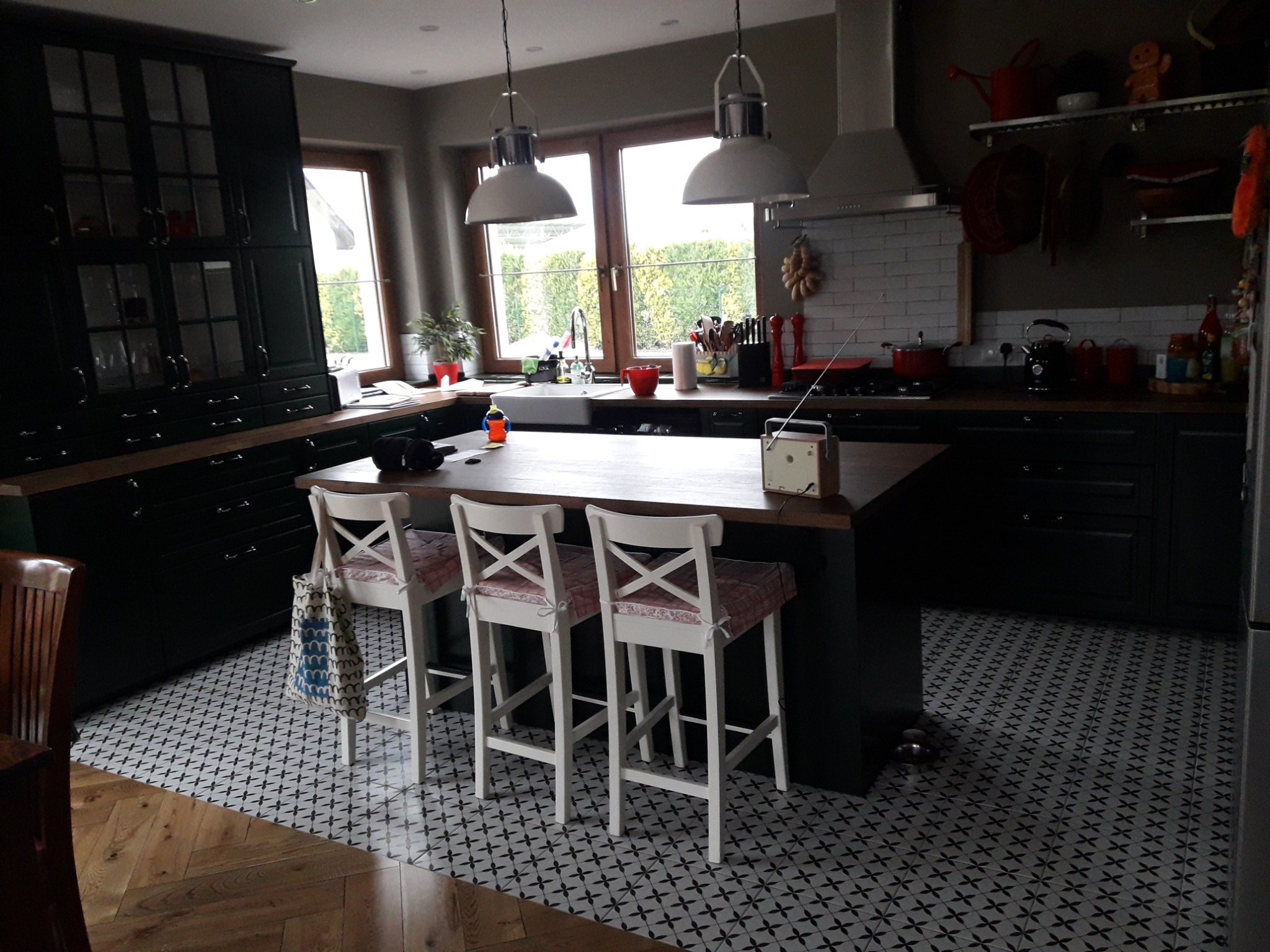 Ciemnozielona kuchnia z wyspą, białymi hokerami z wzorzystymi poduszkami i czarno-białą wzorzystą podłogą.