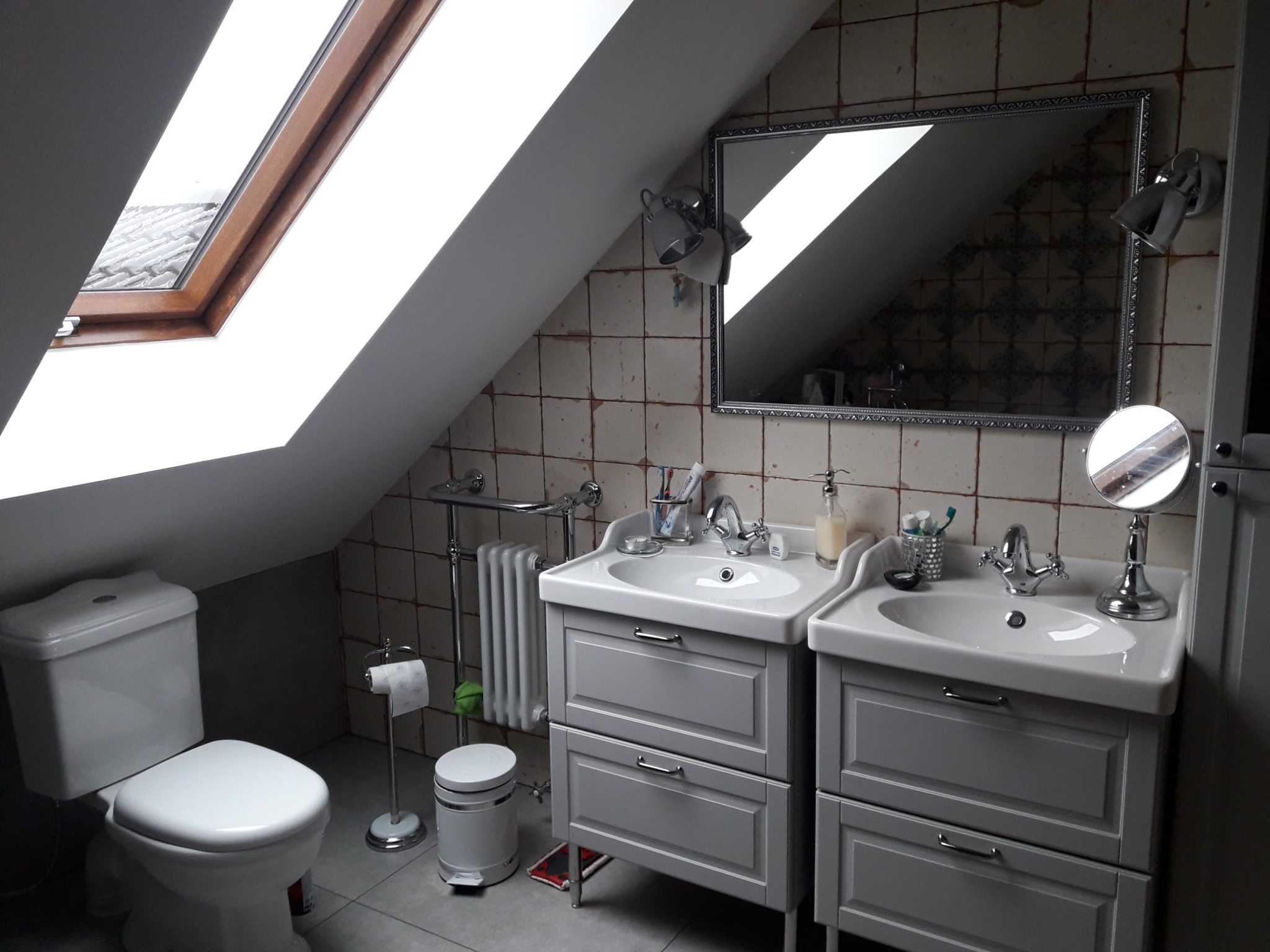 Umywalki z szarymi szafkami w łazience na poddaszu z oknem dachowym i beżowymi płytkami w kratkę.