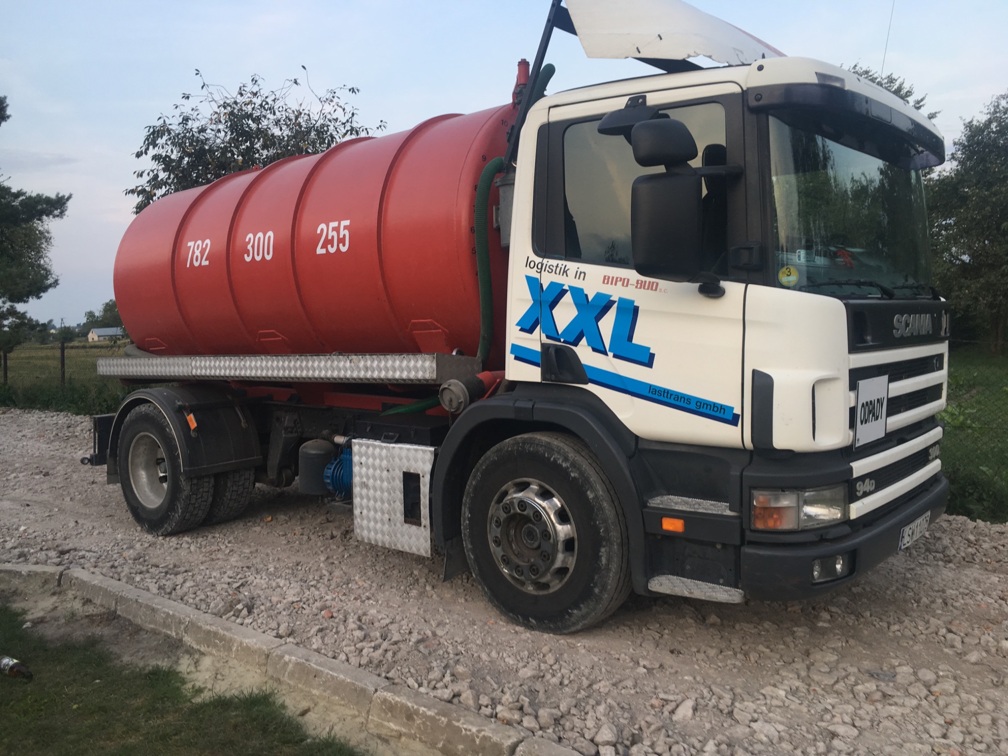 Ciężarówka asenizacyjna Scania 94D z pomarańczową cysterną na gruzowym podłożu, z widocznymi numerami 782, 300, 255 na cysternie i logo firmy logistik in BIPO-BUD S.C. XXL lasttrans gmbh.