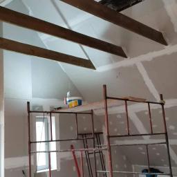 Karol Michałowski - Poddasze w trakcie remontu, widoczne drewniane belki stropowe, ściany pokryte płytami gipsowo-kartonowymi przygotowane do szpachlowania, rusztowanie malarskie i prowizoryczne oświetlenie żarówką.