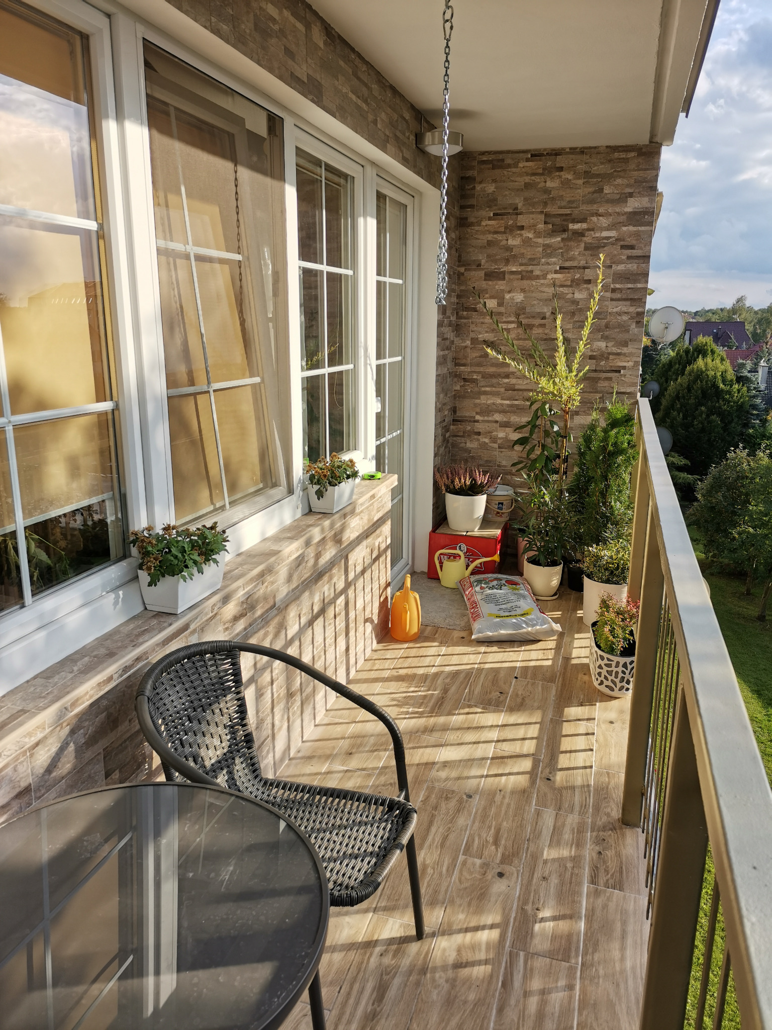 Umeblowany balkon z drewnopodobnymi płytkami, rattanowym krzesłem i szklanym stolikiem. Na ścianie dekoracyjna okładzina imitująca kamień, a na parapecie i podłodze doniczki z roślinami i akcesoria...