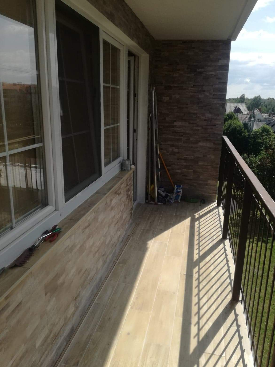 Zabudowany balkon z brązową balustradą, wykończony płytkami imitującymi kamień na ścianach i podłodze, widoczne okno przesuwne i drabina oparta o ścianę.