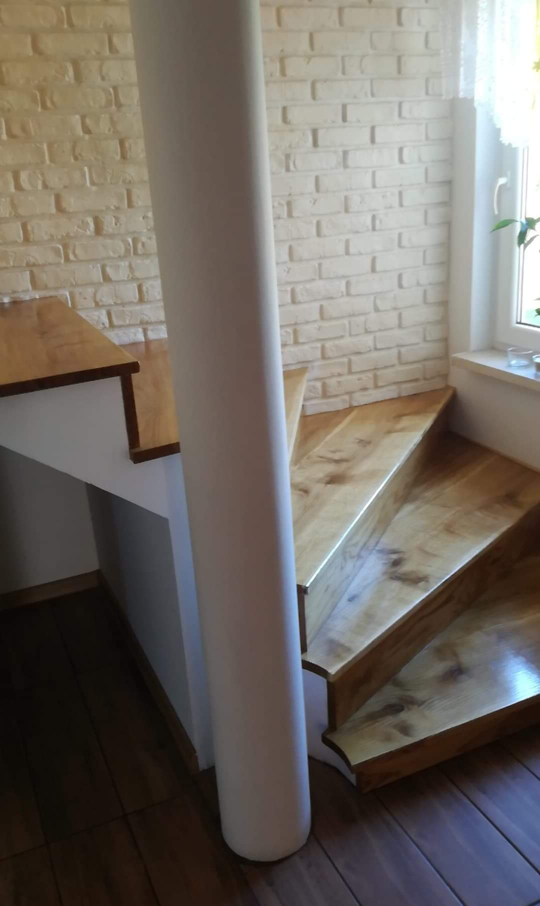 Drewniane schody z jasnymi stopniami i ciemniejszą podłogą, wsparte na białym słupie, z tłem ściany z białej cegły i oknem z firanką w tle.
