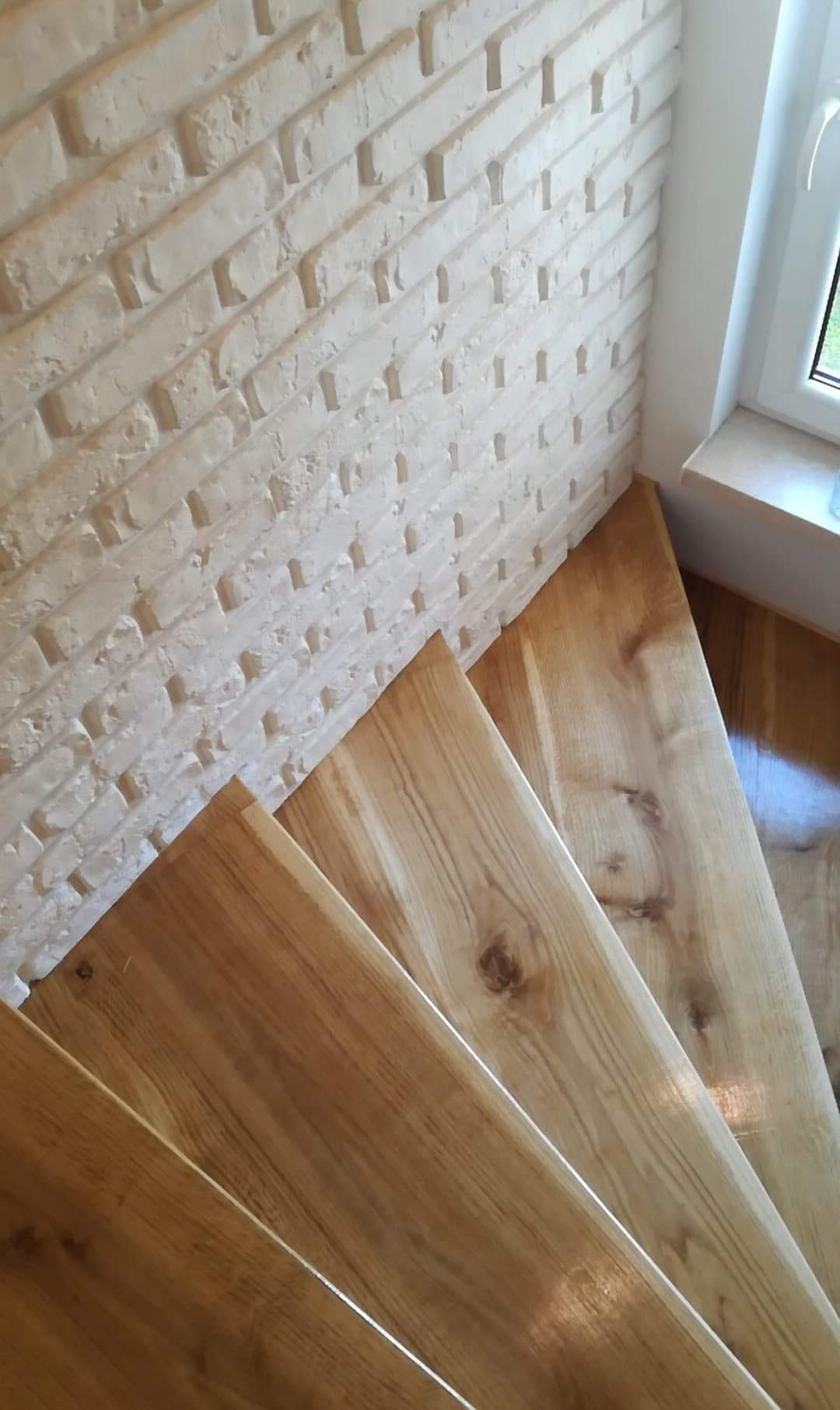 Schody drewniane z naturalnego dębu, widok z góry, z białym murem z cegły dekoracyjnej w tle, oświetlenie dzienne.