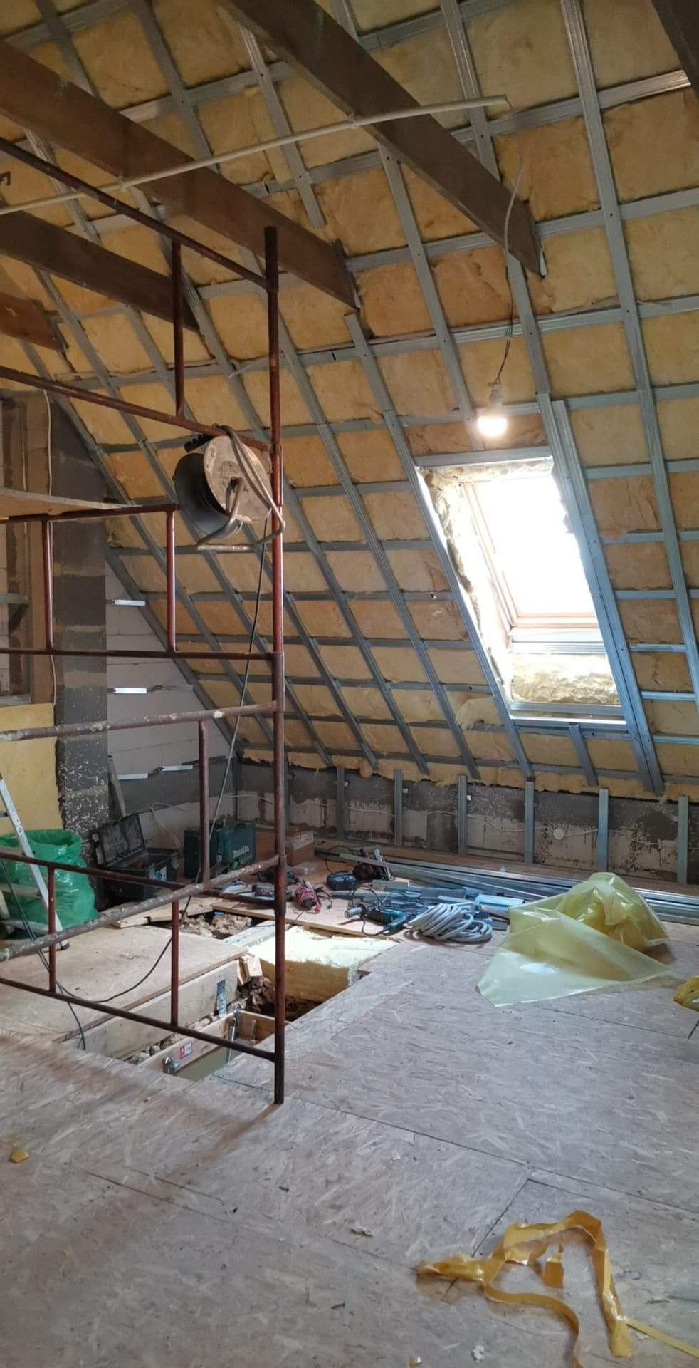 Szkielet metalowej konstrukcji sufitu podwieszanego z izolacją z wełny mineralnej na poddaszu w trakcie montażu w Warszawie, widoczne rusztowanie i narzędzia.