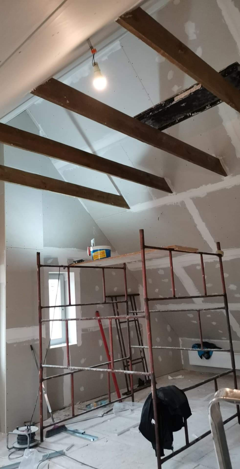 Poddasze w trakcie remontu, widoczne drewniane belki stropowe, ściany pokryte płytami gipsowo-kartonowymi przygotowane do szpachlowania, rusztowanie malarskie i prowizoryczne oświetlenie żarówką.