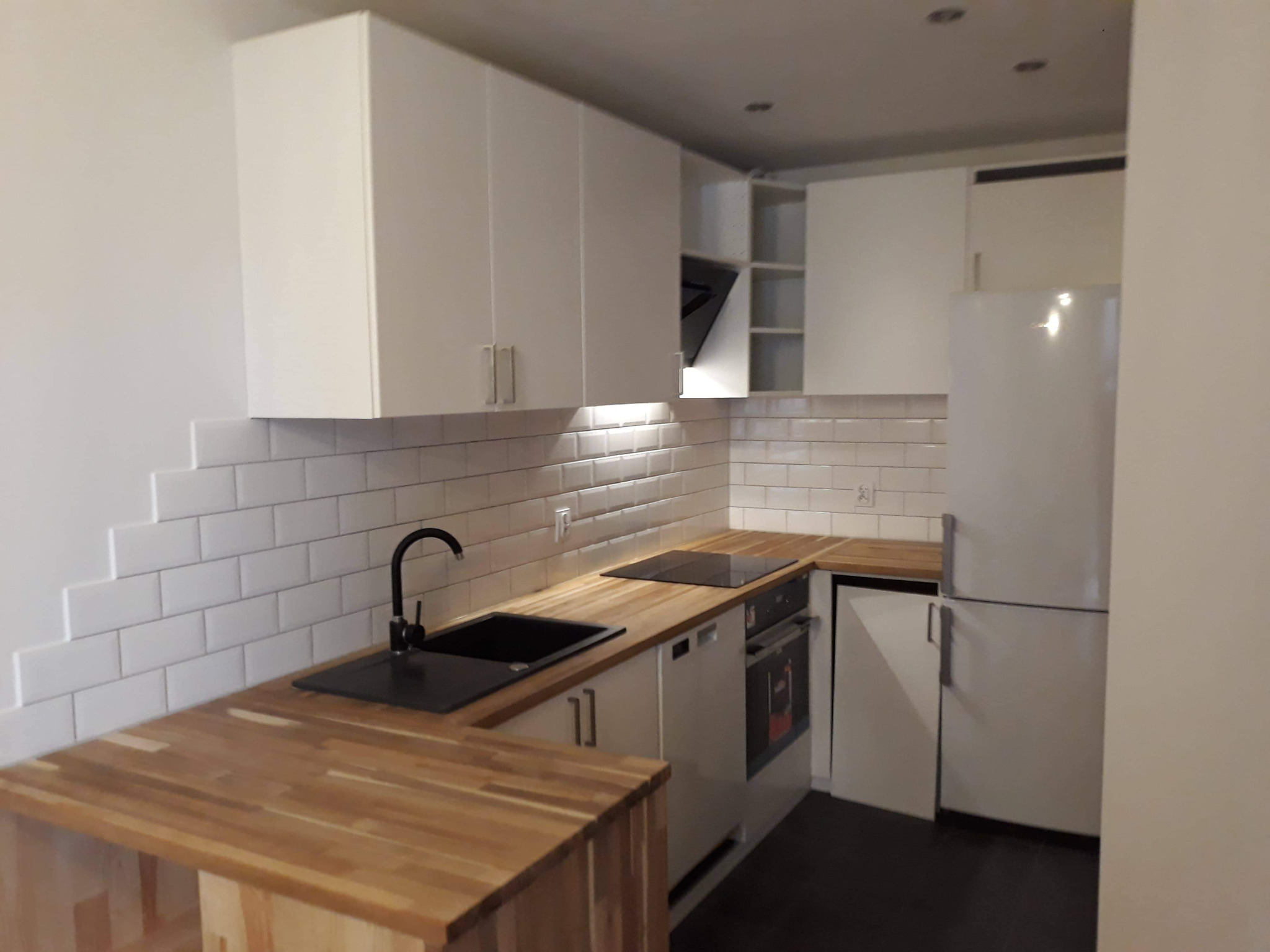 Nowoczesna, jasna kuchnia z białymi szafkami, drewnianym blatem i czarną armaturą, wykończona białymi płytkami ułożonymi schodkowo.