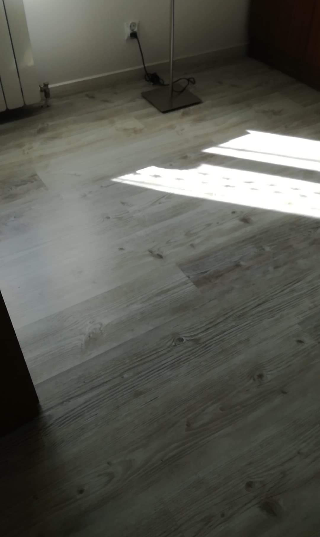 Jasna, drewnopodobna podłoga z paneli, oświetlona promieniami słońca wpadającymi przez okno, widoczna część grzejnika i lampy podłogowej.