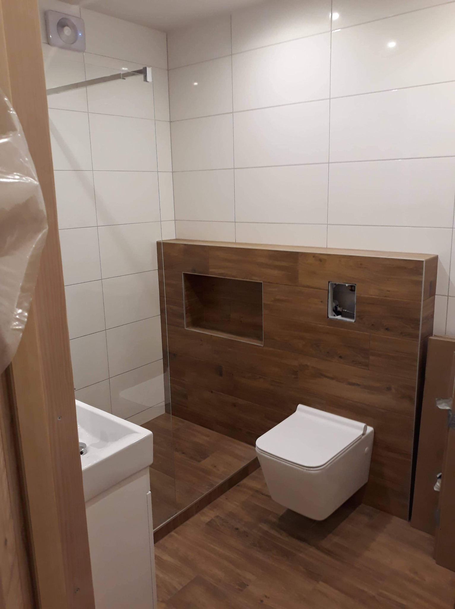 Nowoczesna łazienka z białymi płytkami ściennymi, drewnopodobnymi panelami, wiszącą toaletą i umywalką na szafce, ukazująca minimalistyczny design i funkcjonalne rozwiązania.