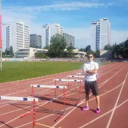 Młody mężczyzna w okularach przeciwsłonecznych, stojący na bieżni lekkoatletycznej z przeszkodami, w tle budynki miejskie i błękitne niebo.