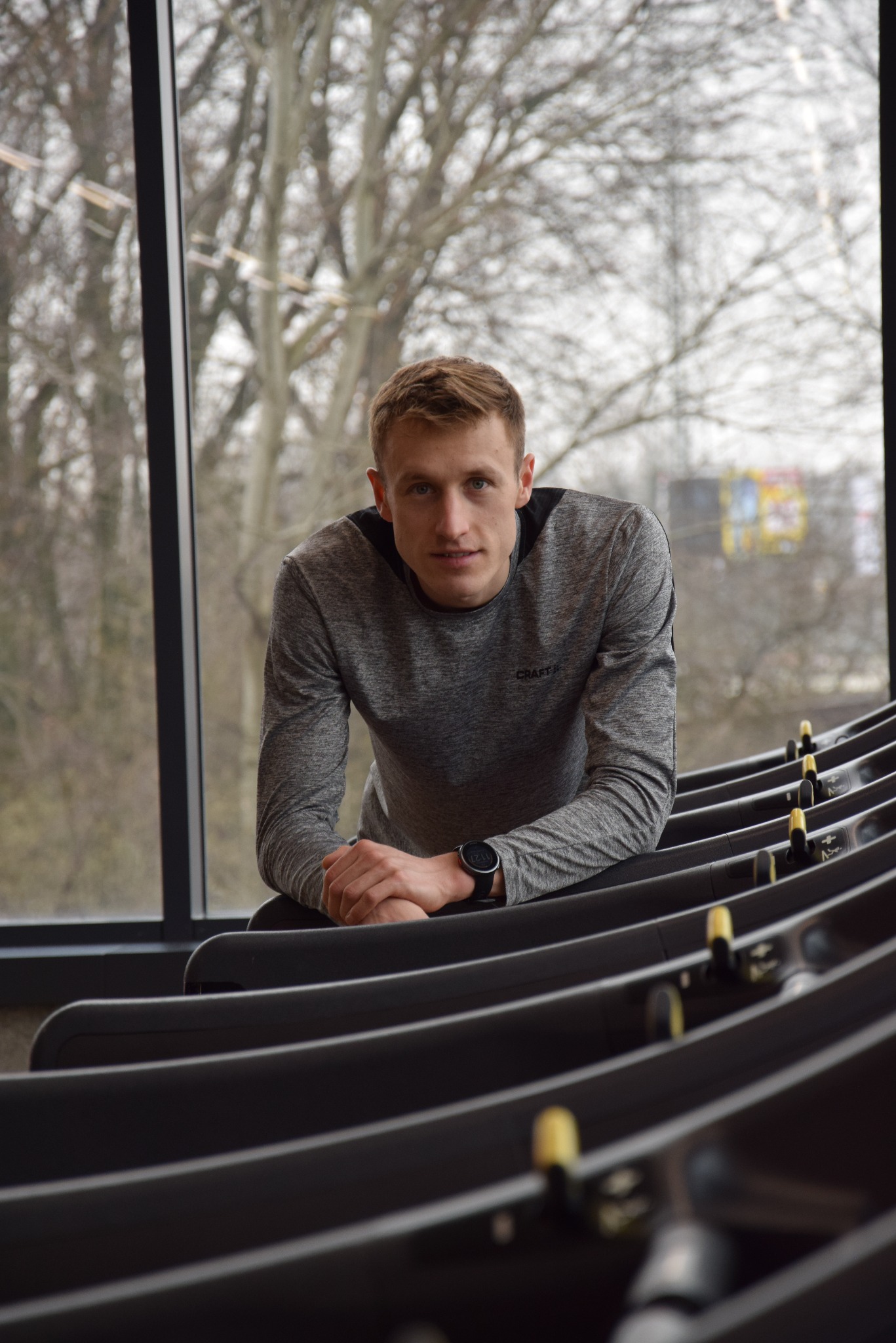 Młody mężczyzna w sportowej koszulce, oparty o bieżnię w siłowni, z widocznym zegarkiem sportowym i dużym oknem w tle. Ujęcie z bliska, naturalne światło.