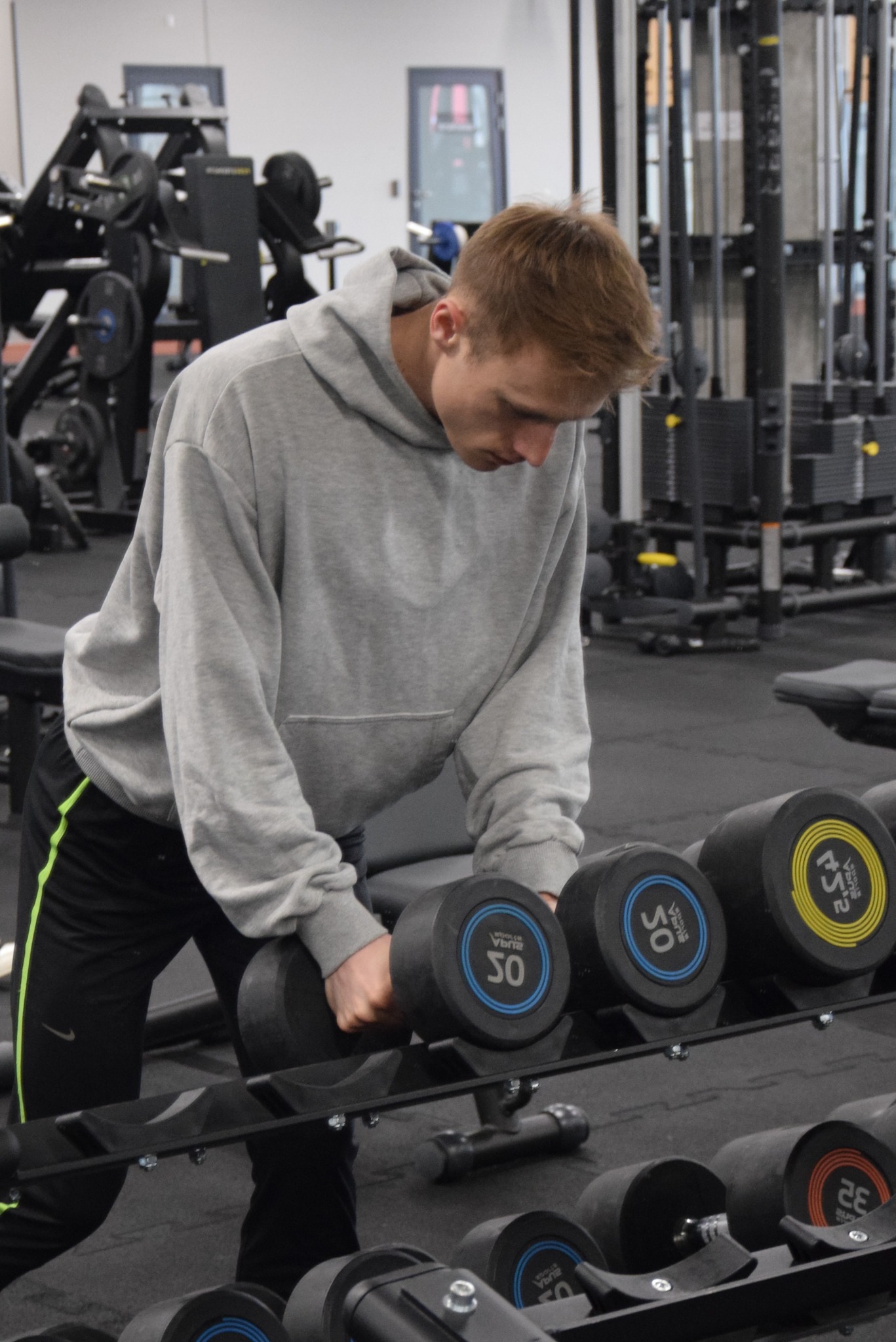 Młody mężczyzna w szarej bluzie z kapturem ćwiczy z hantlami w siłowni. W tle widoczne maszyny do ćwiczeń i ciężary.