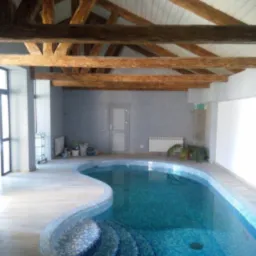 Kryty basen z mozaiką w odcieniach błękitu, z drewnianą konstrukcją dachu i szarą ścianą w tle.