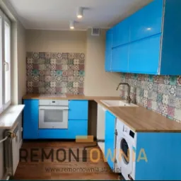 Niebieska zabudowa kuchenna z drewnianym blatem i wzorzystą ścianą nad blatem roboczym, widoczna pralka w zabudowie, oświetlenie punktowe sufitowe.