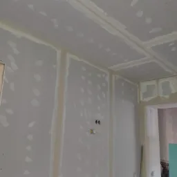 Wnętrze w trakcie prac wykończeniowych: ściany i sufit pokryte płytami kartonowo-gipsowymi, z widocznymi miejscami łączeń i szpachlowania, przygotowane do dalszych etap&oacute;w prac malarskich.