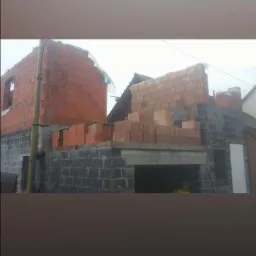 Budowa domu jednorodzinnego w trakcie wznoszenia murów z bloczków betonowych i cegły ceramicznej, widoczny zarys okna i dachu sąsiedniego budynku.