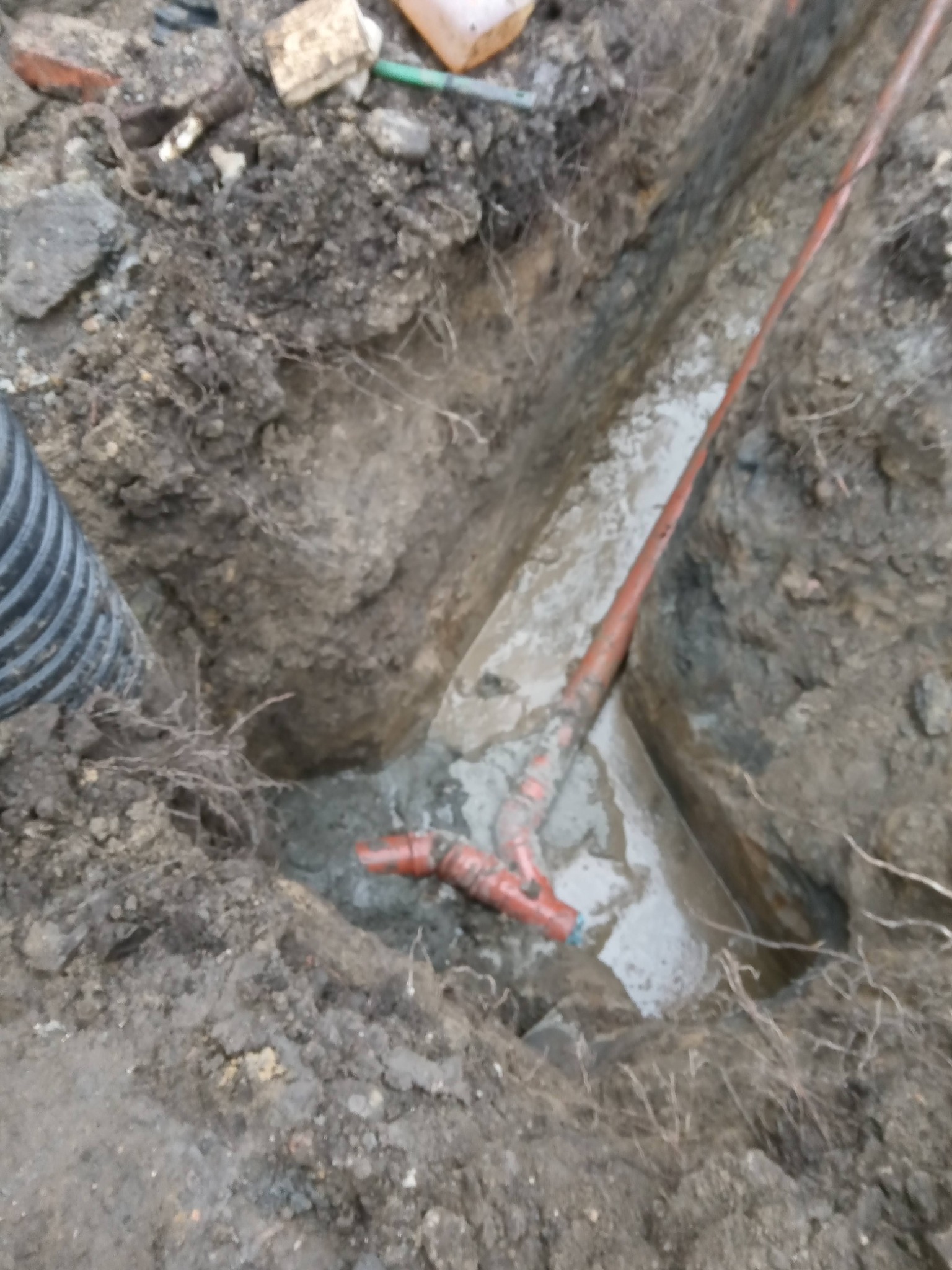 Wykop w ziemi z częściowo ułożonymi rurami kanalizacyjnymi z tworzywa sztucznego w kolorze pomarańczowym, widoczne połączenia i fragment rury karbowanej.